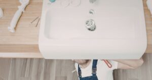 An overhead view of a professional plumber in blue overalls working beneath a large white porcelain sink, with tools and plumbing components laid out on a light wood countertop.