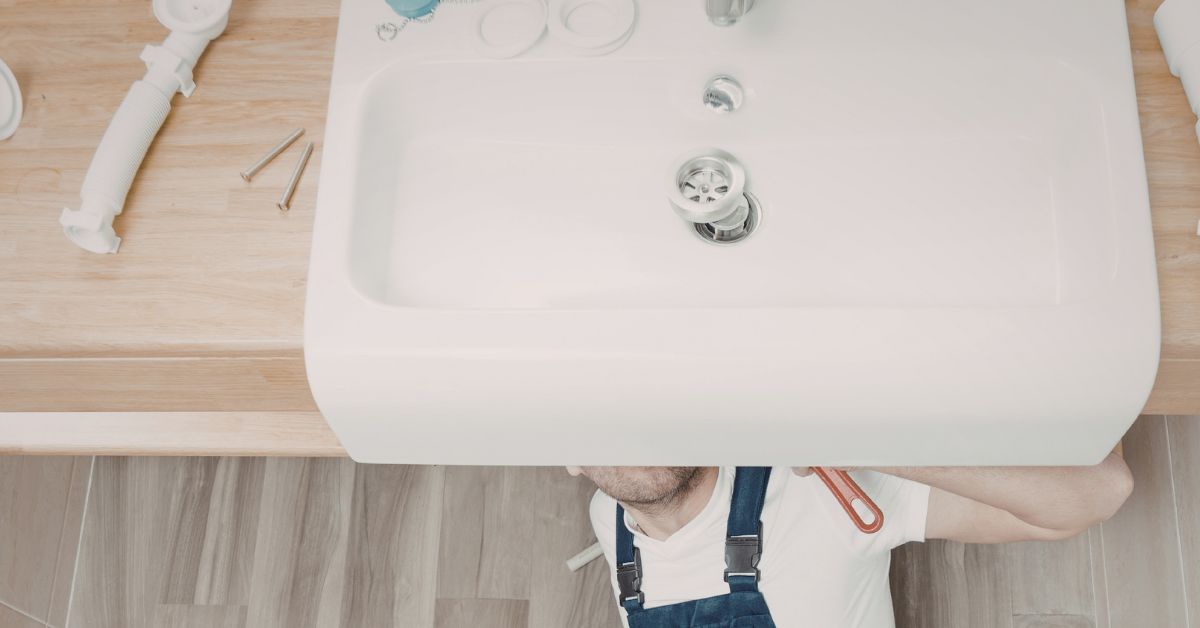 An overhead view of a professional plumber in blue overalls working beneath a large white porcelain sink, with tools and plumbing components laid out on a light wood countertop.