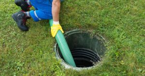 A septic service professional wearing yellow safety gloves guides a large suction hose into a plastic septic riser during a routine maintenance appointment.
