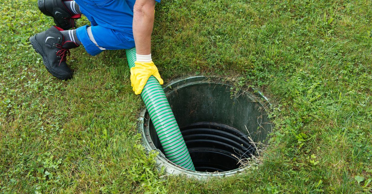 A septic service professional wearing yellow safety gloves guides a large suction hose into a plastic septic riser during a routine maintenance appointment.