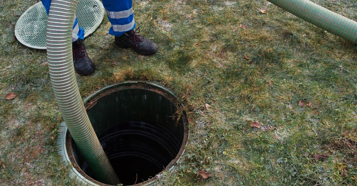 A technician in blue workwear inserts a heavy-duty green suction hose into an open septic tank riser on a residential lawn.